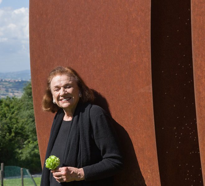 Portrait de l'artiste Beverly Pepper posant fièrement devant une de ses oeuvres.