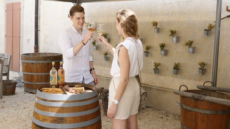 Photo d'un couple trinquant avec des verres de vins dans la cour du Château autour d'un tonneau sur lequel se trouve une planchette du terroir.