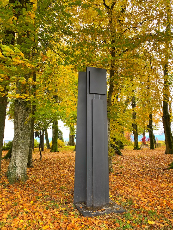 Sculpture totem de couleur noire, de l'artiste Manuel Torres, exposée dans le petit bois du Château de Vullierens. Elle est recouverte de feuilles orangées.