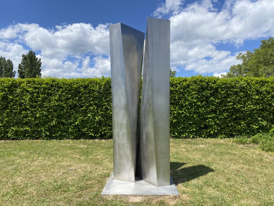 Grand totem métallique de couleur grise exposé en plein air dans le parc de sculptures du Château de Vullierens. L'oeuvre est de Manuel Torres.
