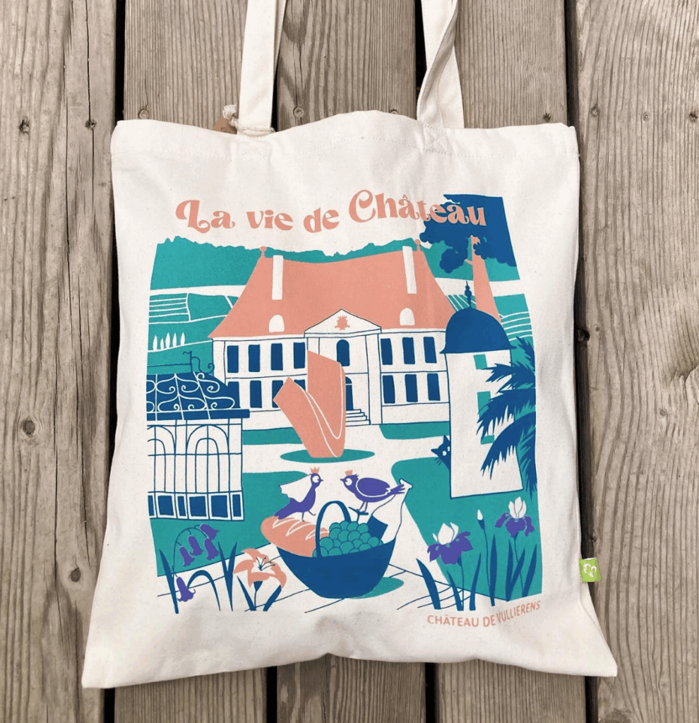 Photo du totbag du Château de Vullierens de couleur beige. L'illustration imprimée dessus représentent les points clés du domaine à savoir le château, des fleurs, la vue sur le lac, les montagnes, le panier pique-nique et les scuptures.